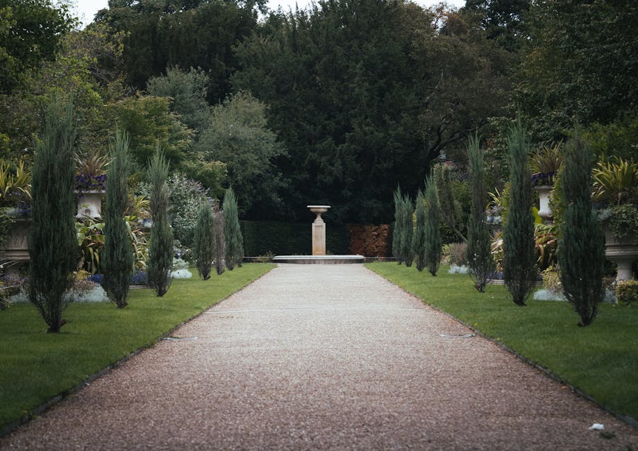 A symmetrical garden pathway lined with neatly trimmed, tall, slender cypress trees on either side, leading towards a classical stone pedestal with a decorative urn at the end of a gravel walkway. The